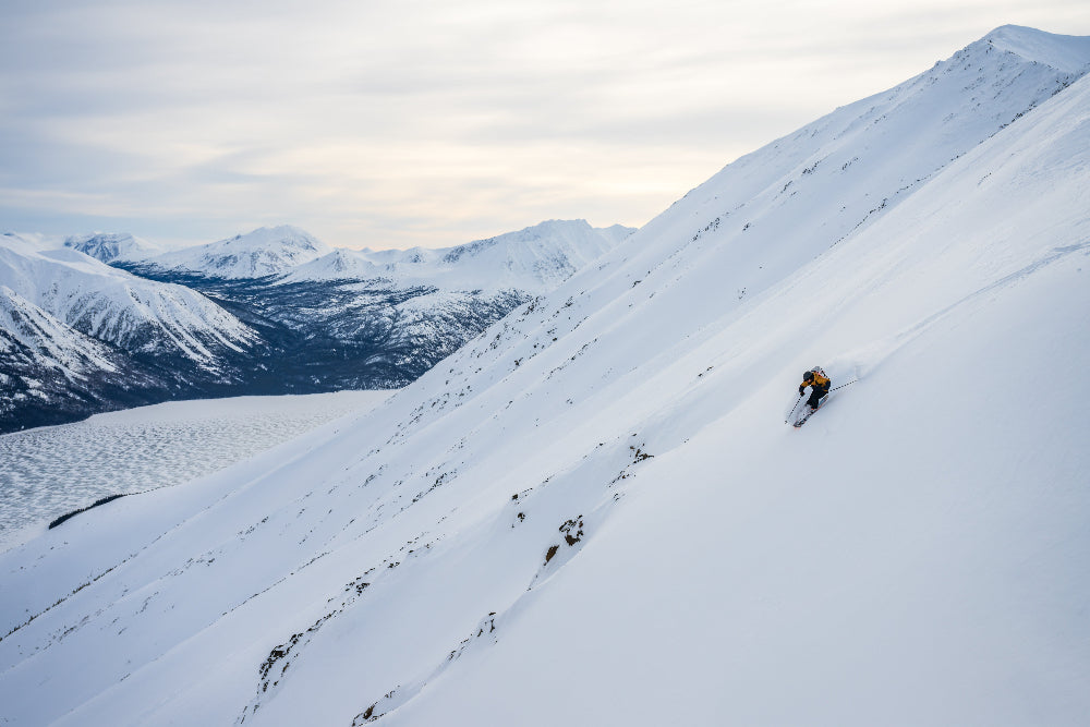 Person backcountry skiing down a snowy mountain slope with a scenic view of other mountains in the background, Yukon Canada