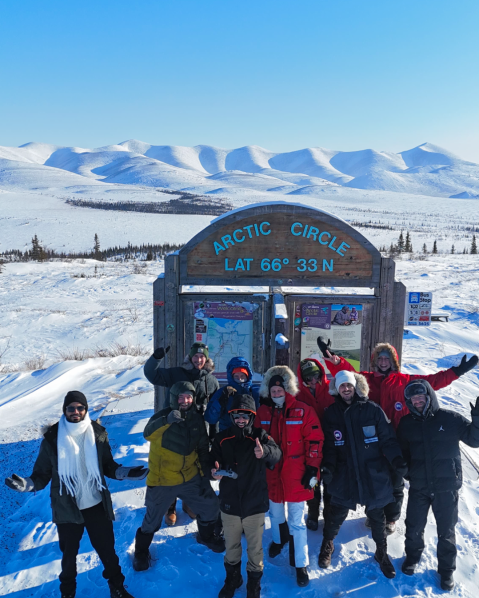Group of people posing in front of an Arctic Circle sign with snowy landscape and mountains in the background.