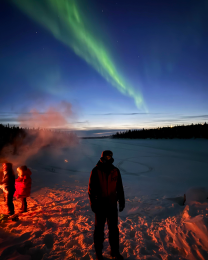 People enjoying the Northern Lights over a snowy landscape with a bonfire.