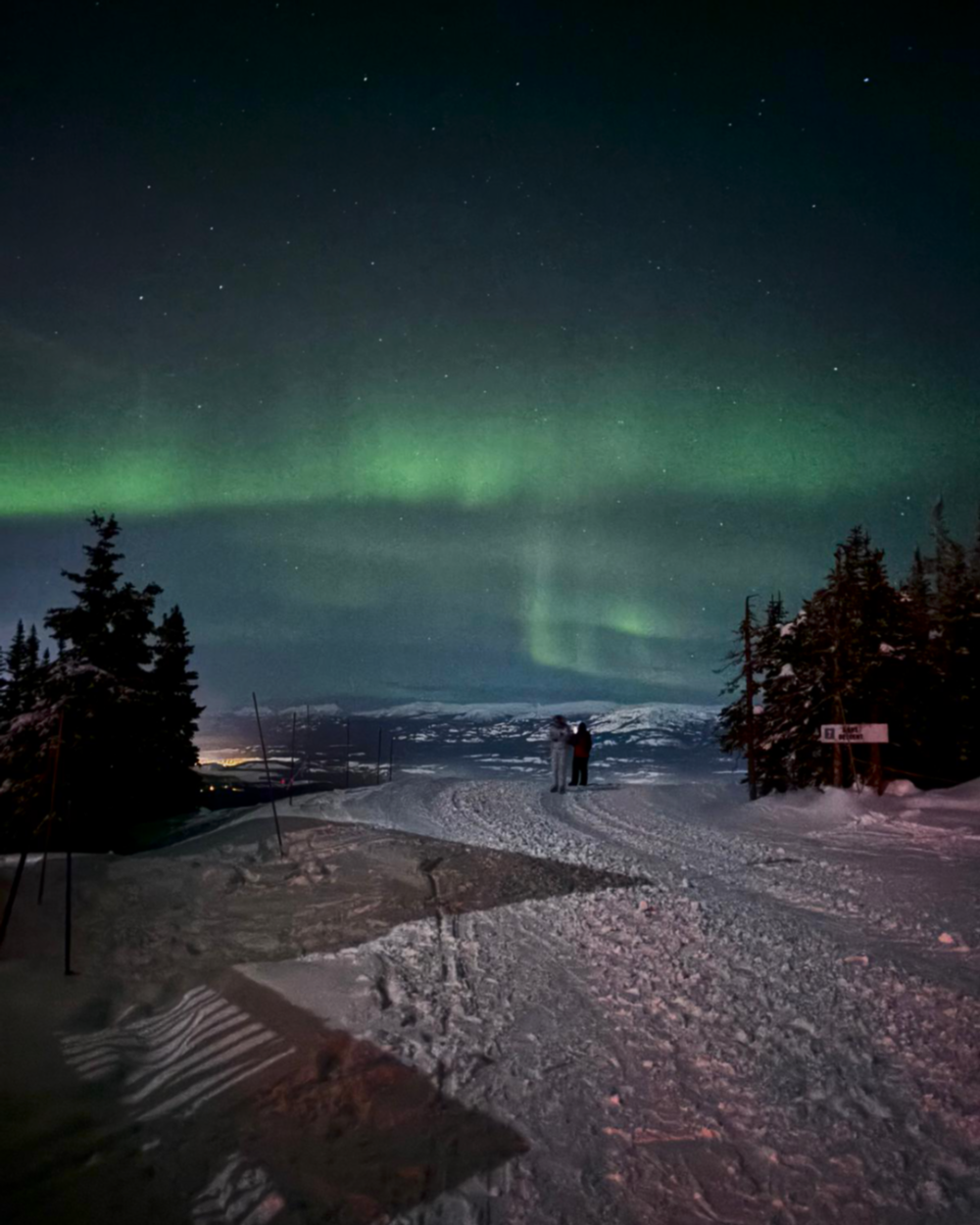 Two people standing on a snow-covered ground with the Aurora Borealis in the sky.
