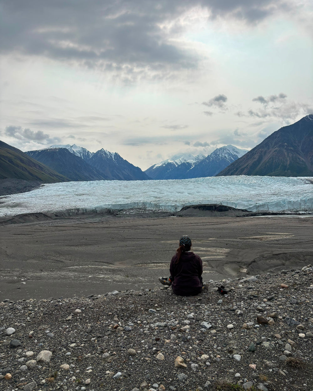 A person sits on a rocky glacier plain facing a large glacier and snow-capped mountains under a cloudy sky. Donjek Glacier, Kluane National Park, Yukon, Canada