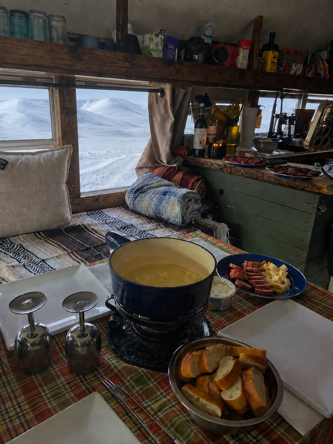 A cozy rustic kitchen scene inside a camper or small cabin, with a blue pot of soup on a burner, bread slices, sausage, and plates on a plaid tablecloth. A large window shows a snowy mountain landscape outside.