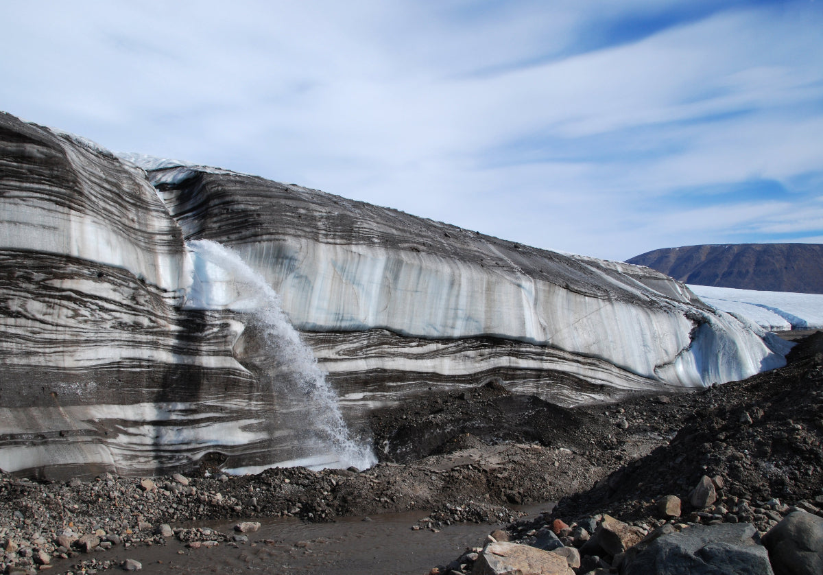 Glacier with visible layers and a small waterfall, set against a blue sky.