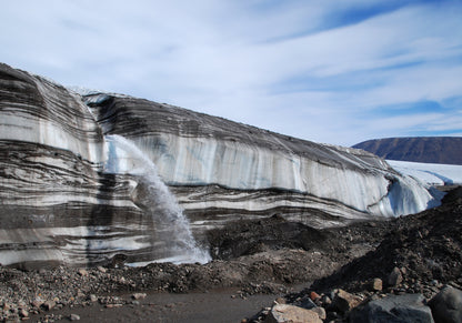 Glacier with visible layers and a small waterfall, set against a blue sky.