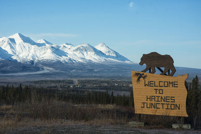 A wooden 'Welcome to Haines Junction' sign with a bear silhouette stands in a landscape with snow-covered mountains, evergreen trees, and a clear blue sky.