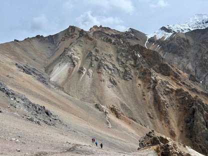 A wide mountain landscape with rocky tan and brown slopes, sparse clouds, and snow patches on distant peaks. A few tiny hikers appear near the bottom center to show scale.