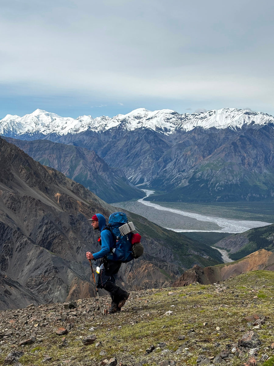 A hiker with a large blue backpack walks along a rocky trail in a mountainous landscape with snow-capped peaks and a river valley in the background. Yusquatch ACMG certified guide hiking Donjek route, Kluane Yukon, Canada