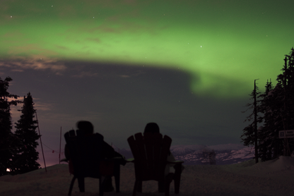 Two people sitting in wooden chairs under the Northern Lights, in Whitehorse Yukon