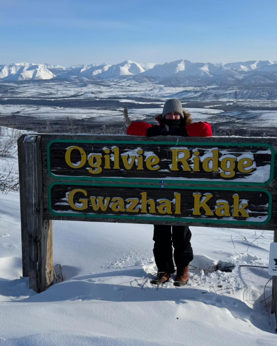 Person standing behind a sign with 'Ogilvie Ridge Gwazhal Kak' in a snowy landscape with mountains.