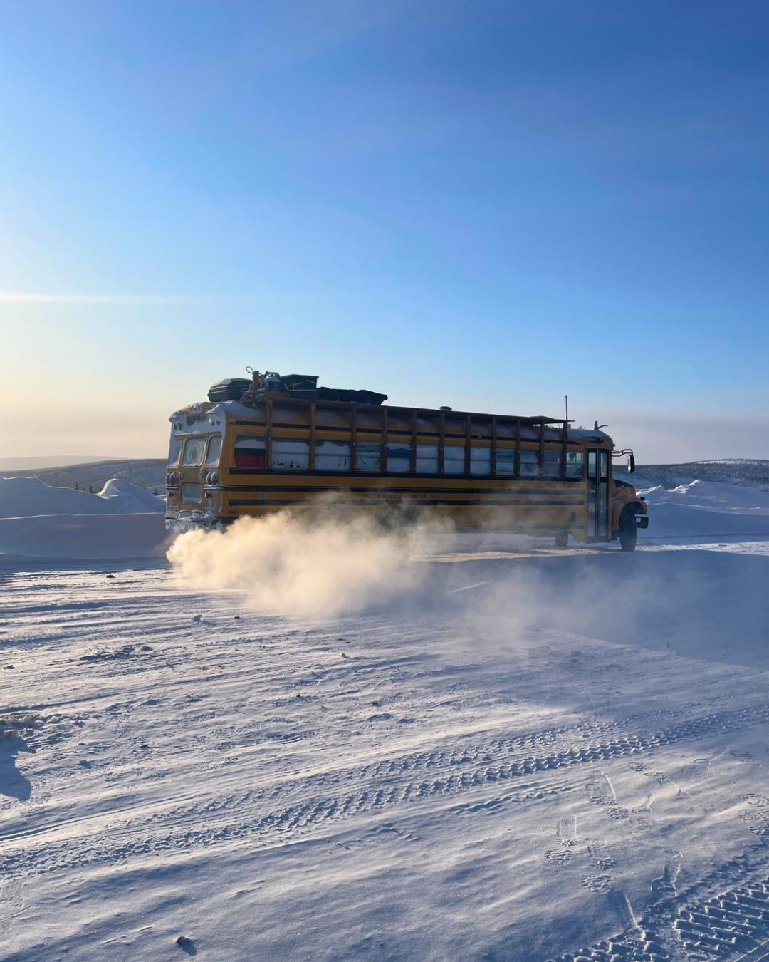 A yellow school bus drives across a snowy landscape, kicking up a cloud of snow in an open, windswept field under a clear blue sky.