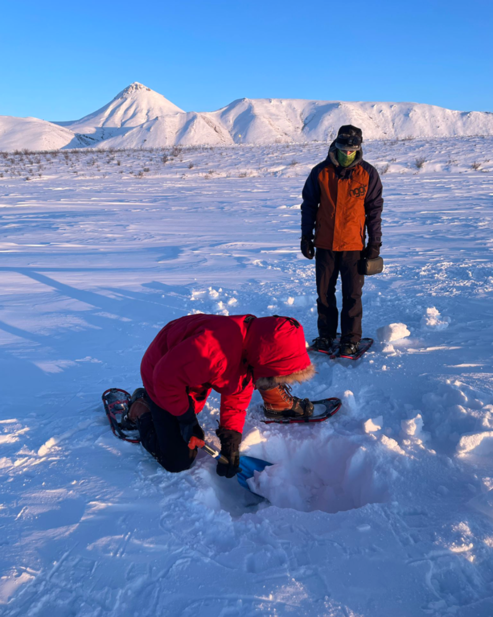 Two people in winter clothing preparing to ice fish on a frozen lake with snow-covered mountains in the background.