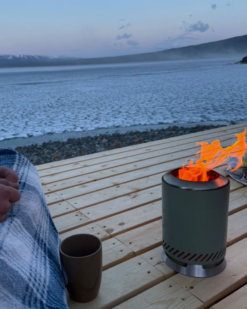 Person sitting by a fire pit on a wooden deck overlooking a lake with mountains in the background. Rooftop patio with cozy campire on top.